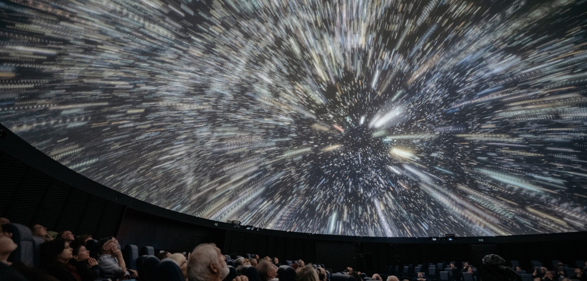 Menschen sitzen in einem Planetarium und blicken auf eine Kuppel, die eine dynamische Animation mit Lichtspuren und Sternen zeigt (ChatGPT).
