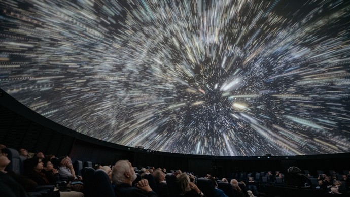 Menschen sitzen in einem Planetarium und blicken auf eine Kuppel, die eine dynamische Animation mit Lichtspuren und Sternen zeigt (ChatGPT).