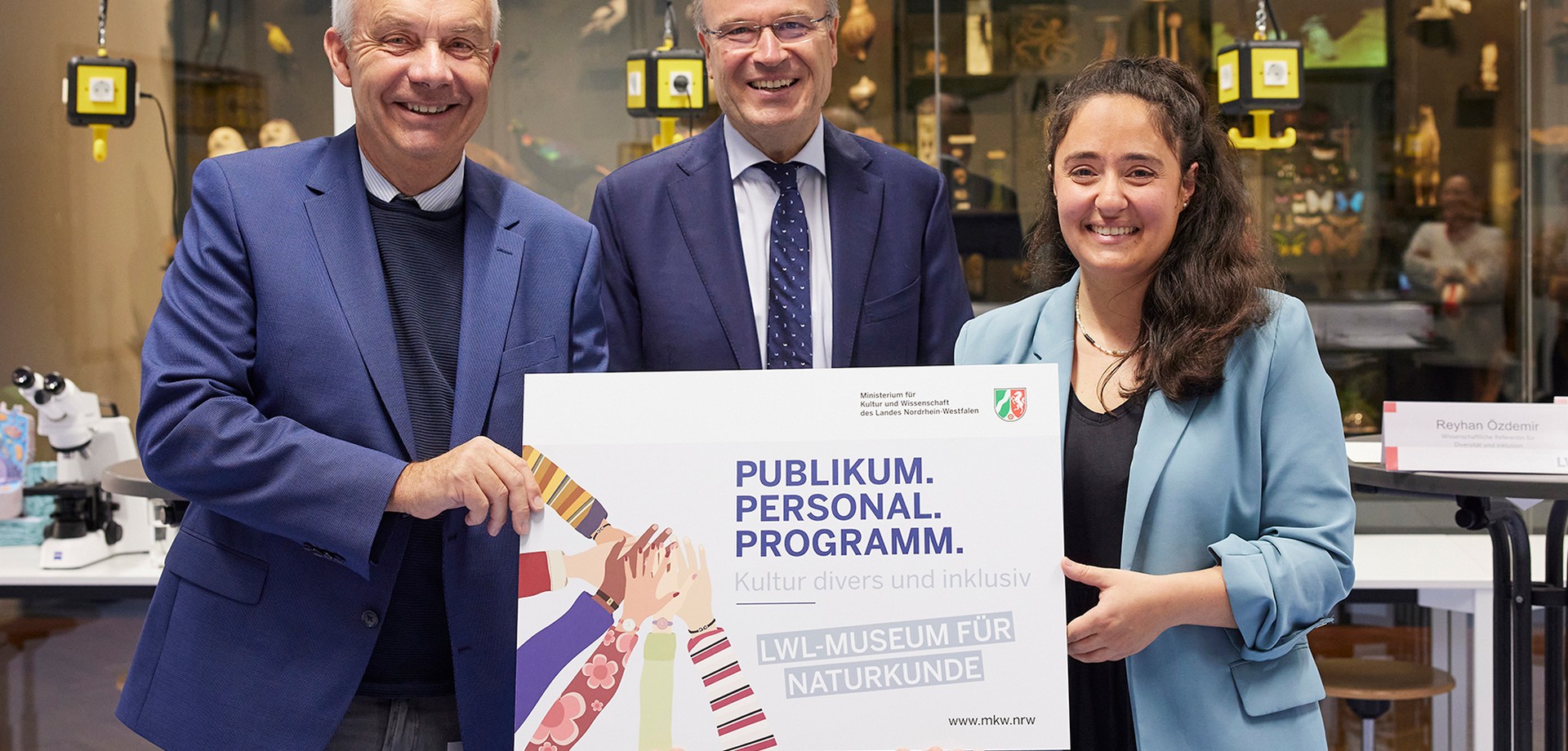 Drei Personen präsentieren ein Plakat mit dem Text ‚Publikum. Personal. Programm. Kultur divers und inklusiv – LWL-Museum für Naturkunde'. (ChatGPT)