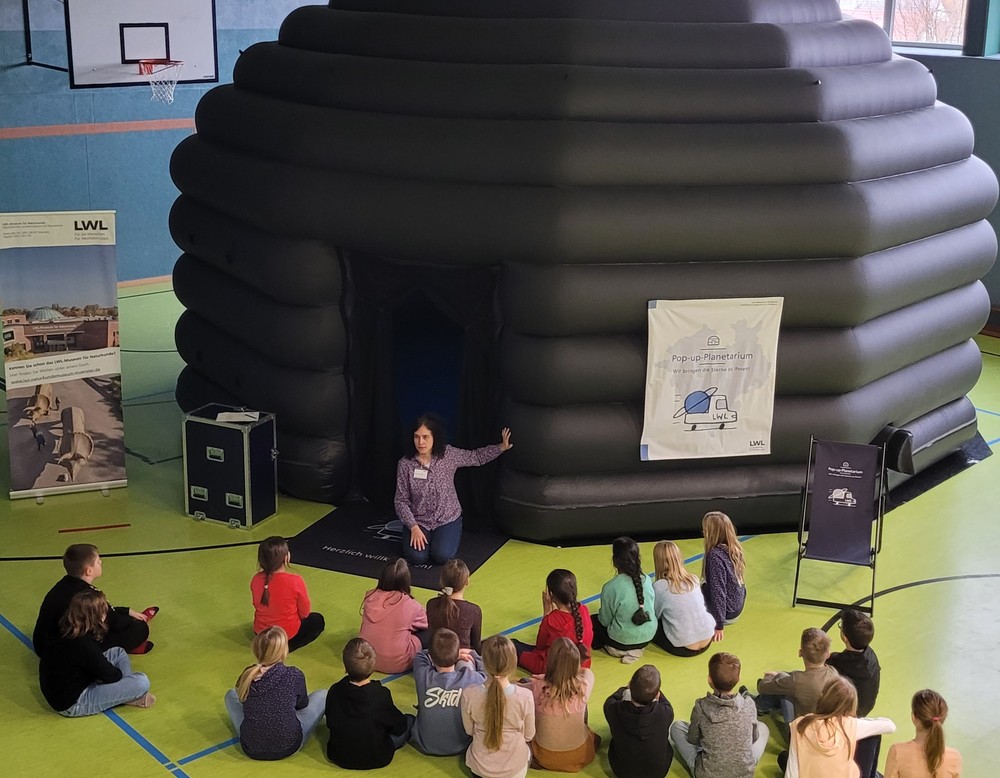 Eine Schulklasse sitzt vor dem Eingang des Pop-up-Planetariums während eine Lehrende Person erklärt.