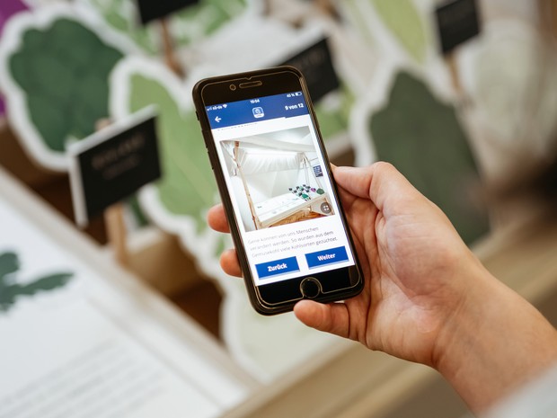 Eine Hand mit einem Handy vor einer Inszenierung eines Gemüsestandes in einer Ausstellung im Museum. Auf dem Handy sind Informationen zur Ausstellung. Eine Hand mit einem Handy vor einer Inszenierung eines Gemüsestandes in einer Ausstellung im Museum. Auf dem Handy sind Informationen zur Ausstellung.
