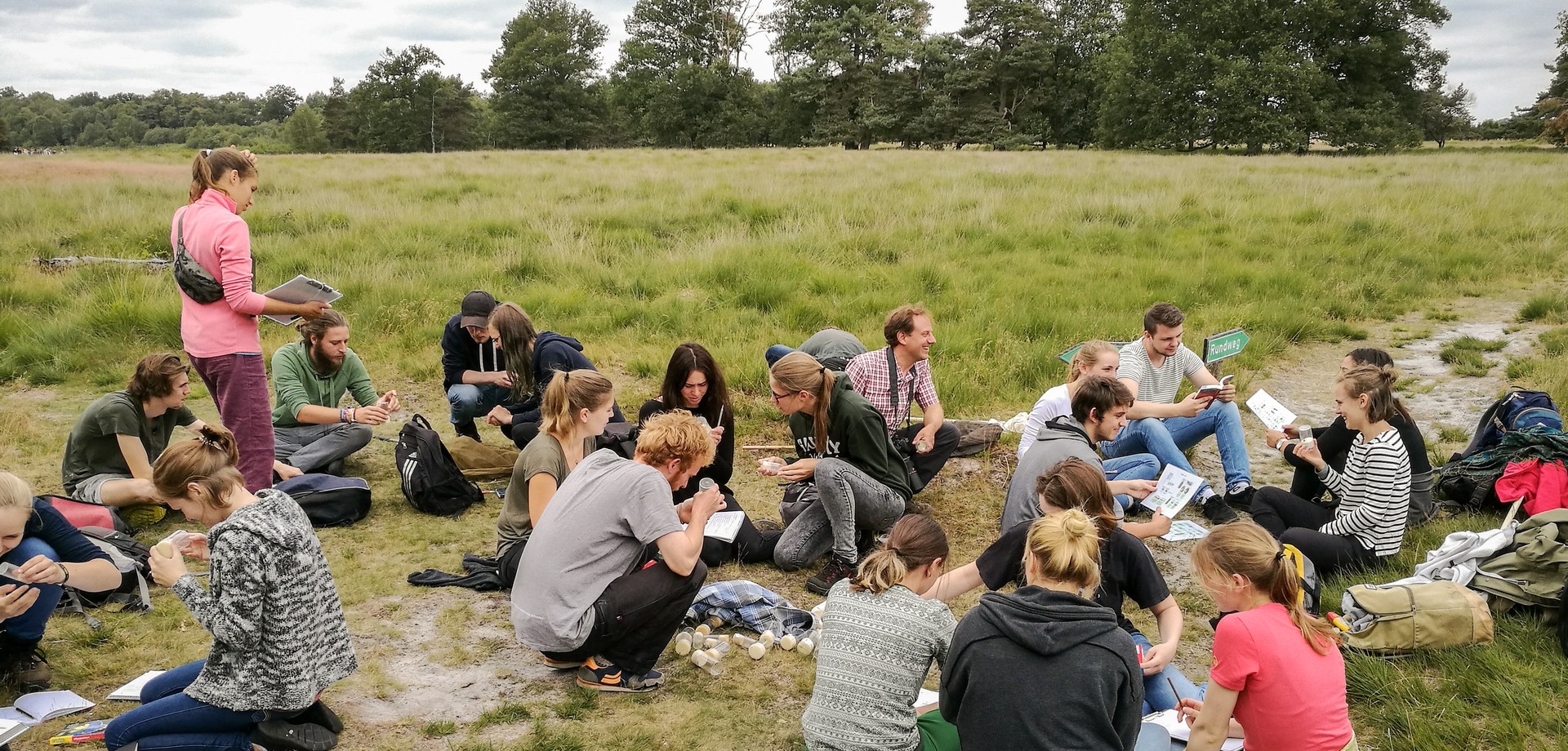 Viele junge Menschen sitzen, hocken und stehen zum Teil mit Büchern, Lupen oder anderen Materialien in den Händen auf einer Wiese. Foto: LWL/Oblonczyk