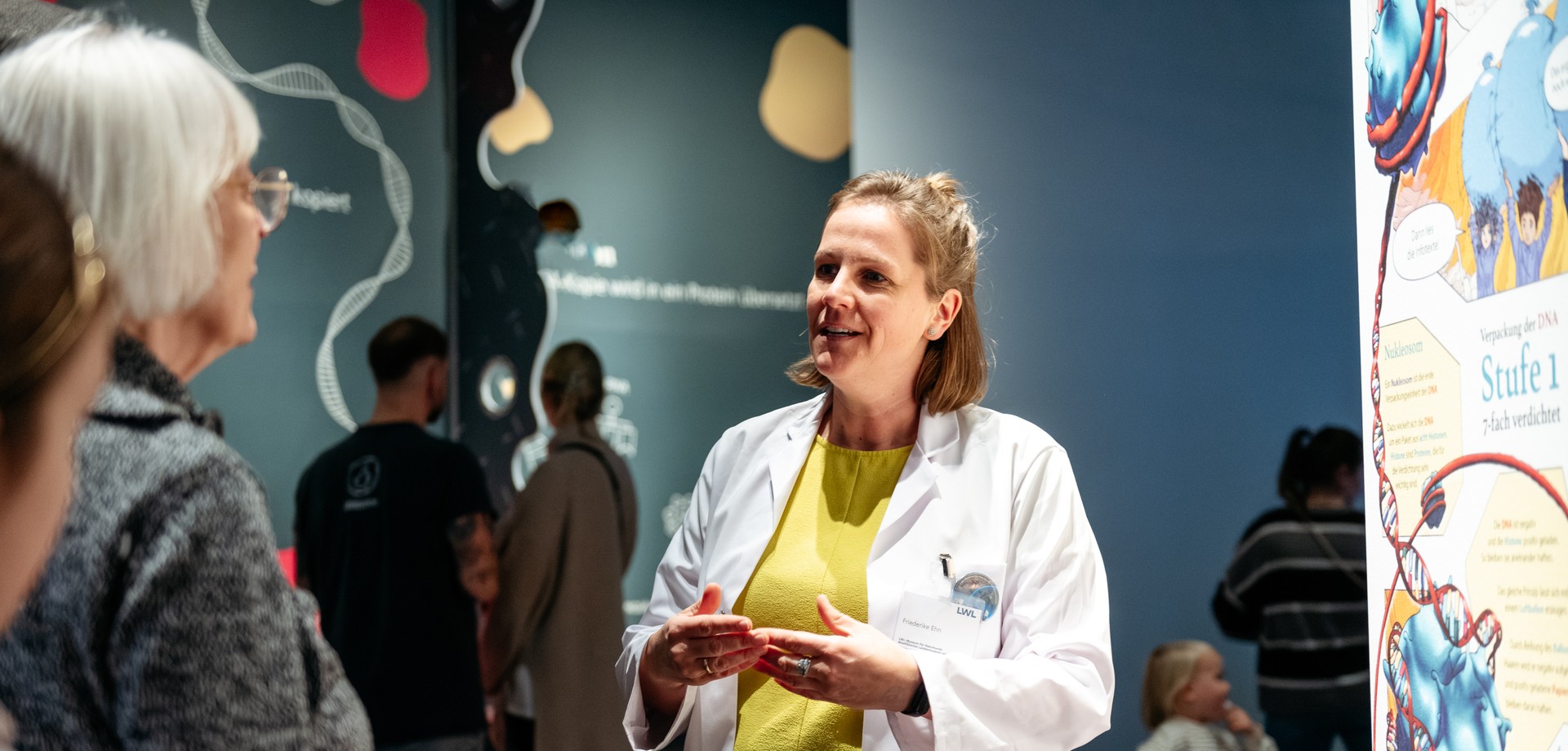 Eine Frau im weißen Laborkittel steht in der Ausstellung und erklärt einer Gruppe etwas.