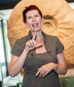 Eine Frau steht vor einem großen Fossil und gibt eine Führung in Gebärdensprache.