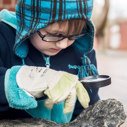Ein Kind mit Arbeitshandschuhen schaut mti einer Lupe auf ein Fossil.