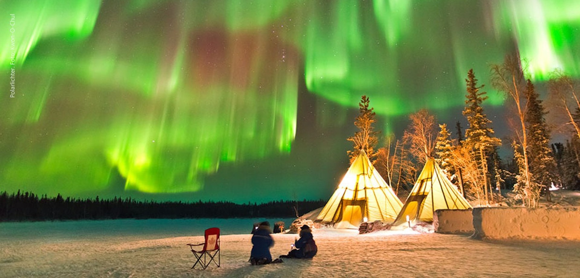 Zwei Personen sitzen vor zwei Zelten im Schnee und beobachten Polarlichter.