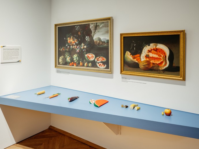 Tisch mit verschiedenen nachgebildeten Obst- und Gemüsesortren zum Ertasten. An der Wand hängen zwei Ölgemälde, die Stillleben von Obst zeigen. (vergrößerte Bildansicht wird geöffnet)