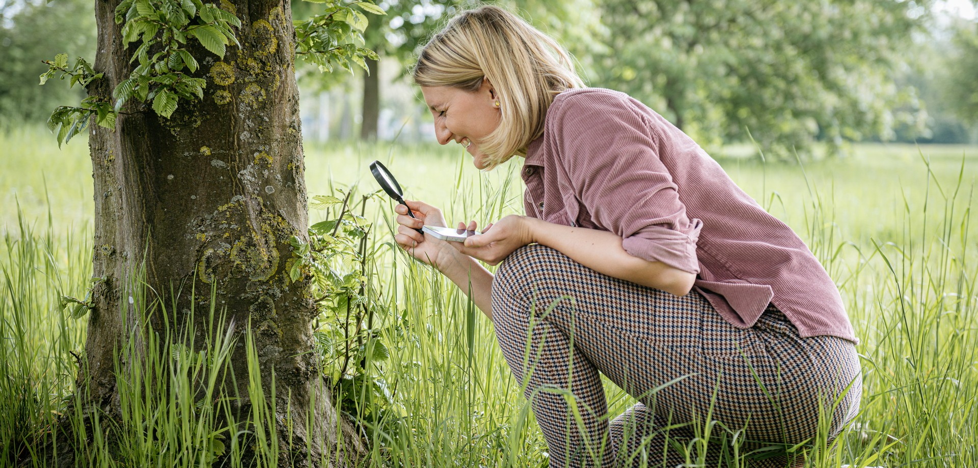 Ein junge Frau mit Lupe und Handy in der Hand schaut sich einen Baum und Pflanzen genauer an.