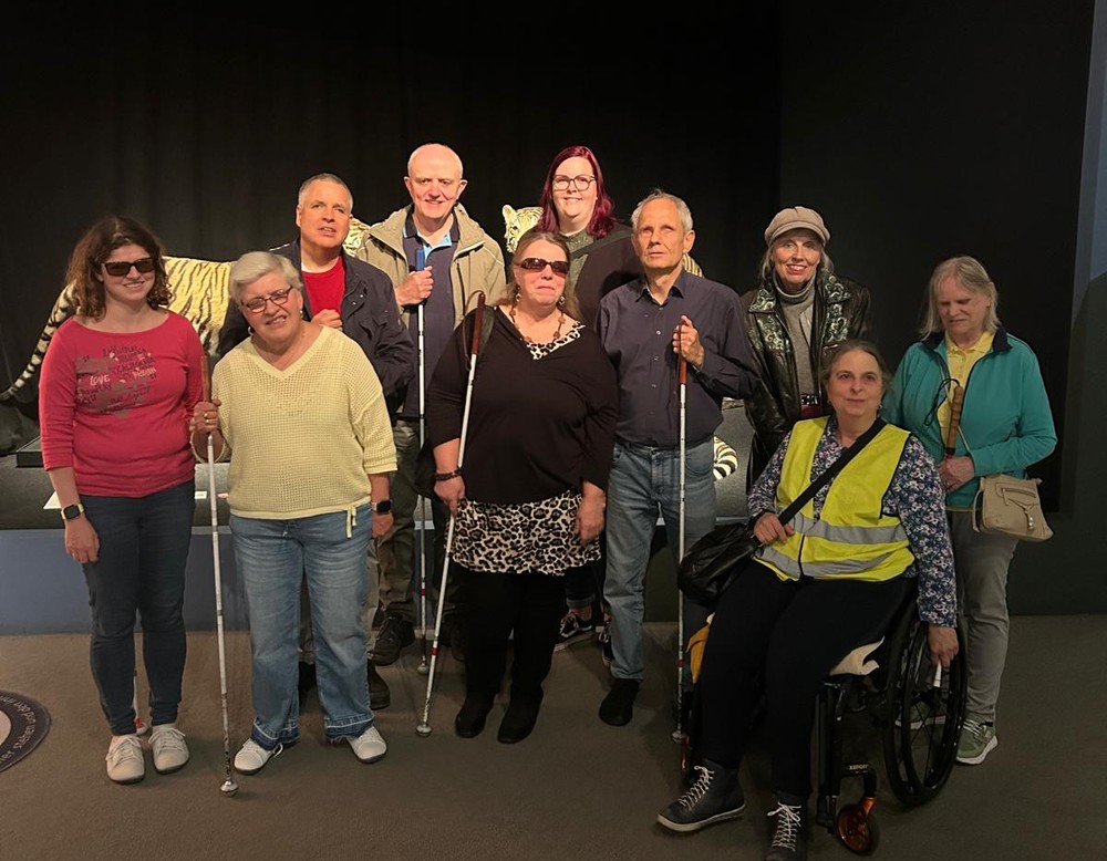 Gruppenfoto mit 10 Mitgliedern des Blinden- und Sehbehindertenvereins Münster vor dem Eingang der Gene-Ausstellung.