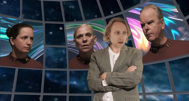 Ein Mann steht mit verschränkten Armen im Vordergrund. Im Hintergrund blicken Gesichter aus Bildschirmen auf ihn herunter.