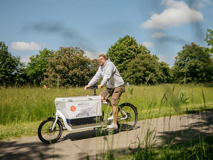 Mann fährt mit einem E-Lastenrad mit der Aufschrift "VELOciraptor" auf einem Radweg durchs Grüne. (vergrößerte Bildansicht wird geöffnet)