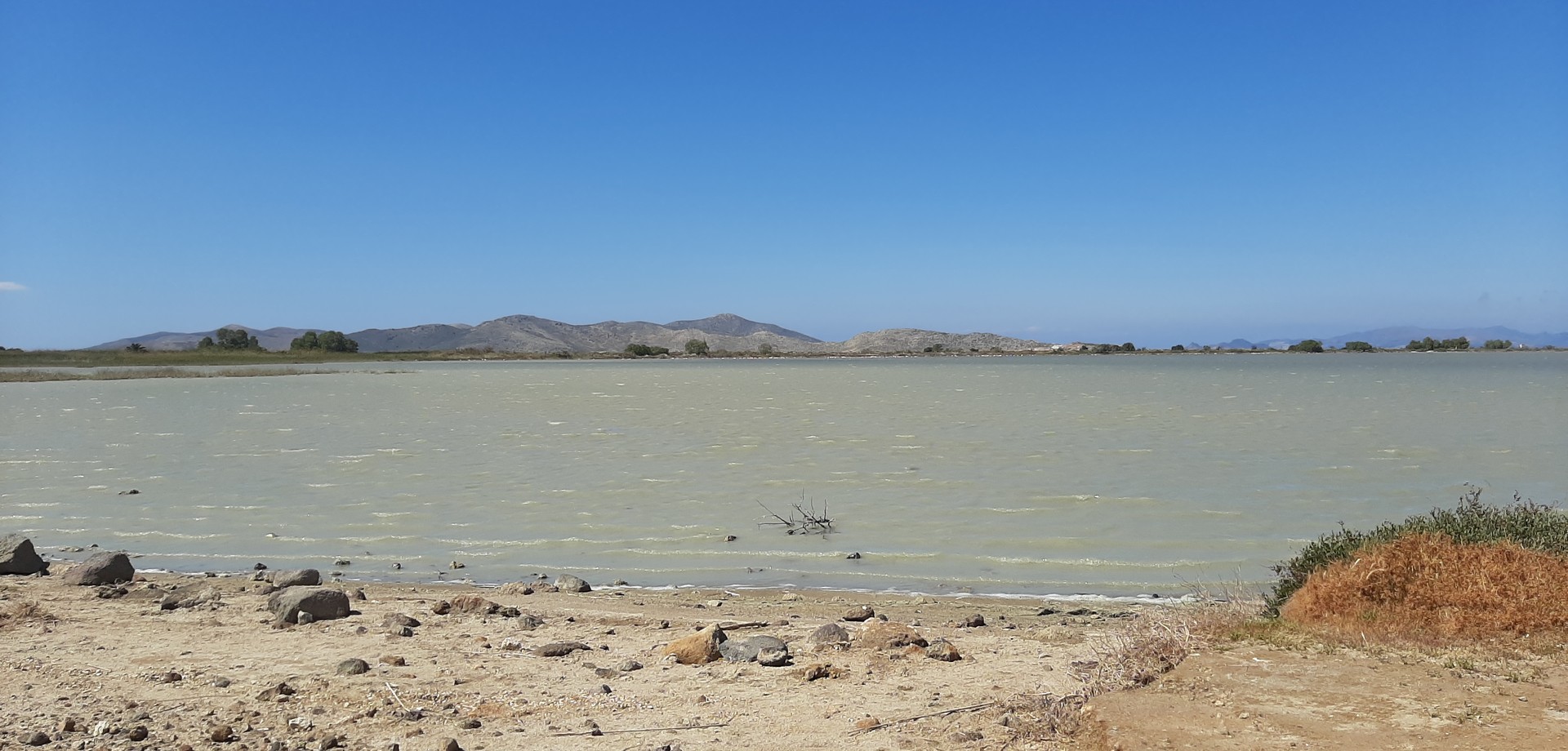 Ein Kalksee mit steinigem Ufer und Bergen am Horizont. Darüber strahlend blauer Himmel.