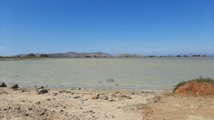 Ein Kalksee mit steinigem Ufer und Bergen am Horizont. Darüber strahlend blauer Himmel.