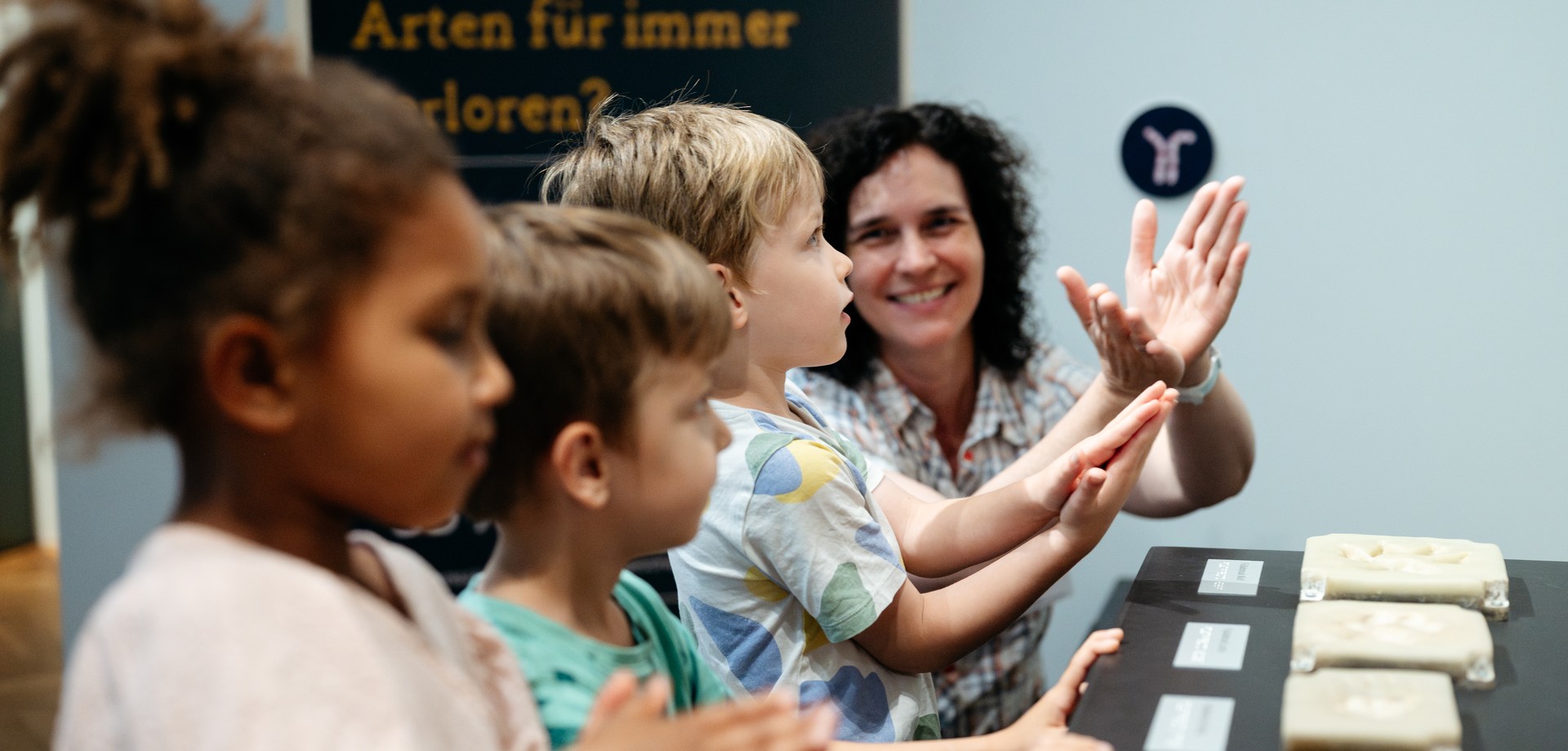 In einer Reihe stehen ein Mädchen, dann zwei Jungen und zuletzt die Museumspädagogin, die etwas erklärt, an einer Taststation.