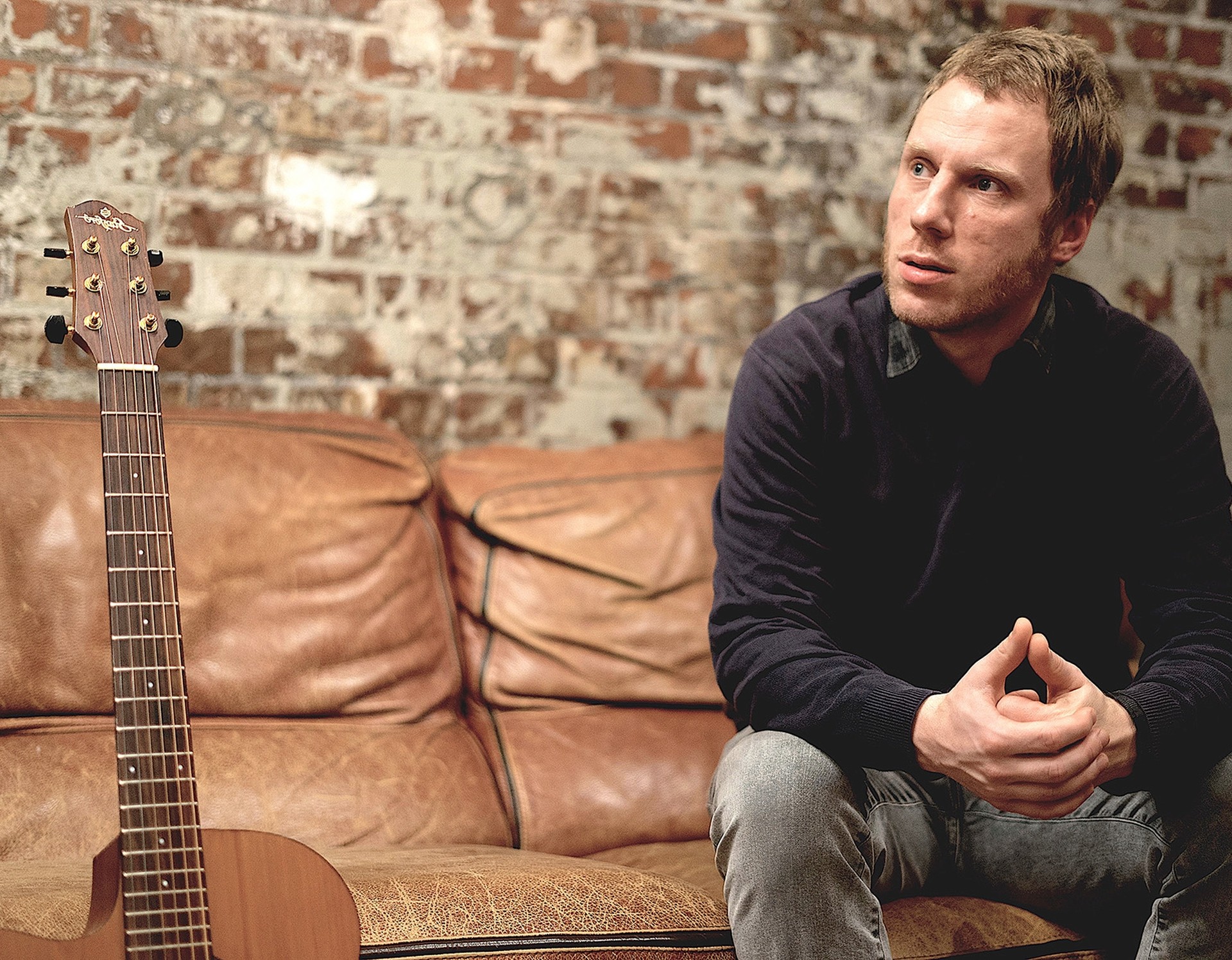 Der Musiker und Komponist Chris Hanson sitzt neben einer Gitarre auf einem Sofa.