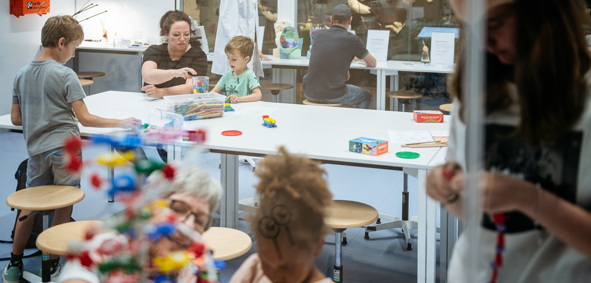 Im Gene-Lab: Mehre Erwachsene sitzen zusammen mit Kindern, schauen durch Mikroskope, bauen DNA-Modelle oder Zellen aus Lego.