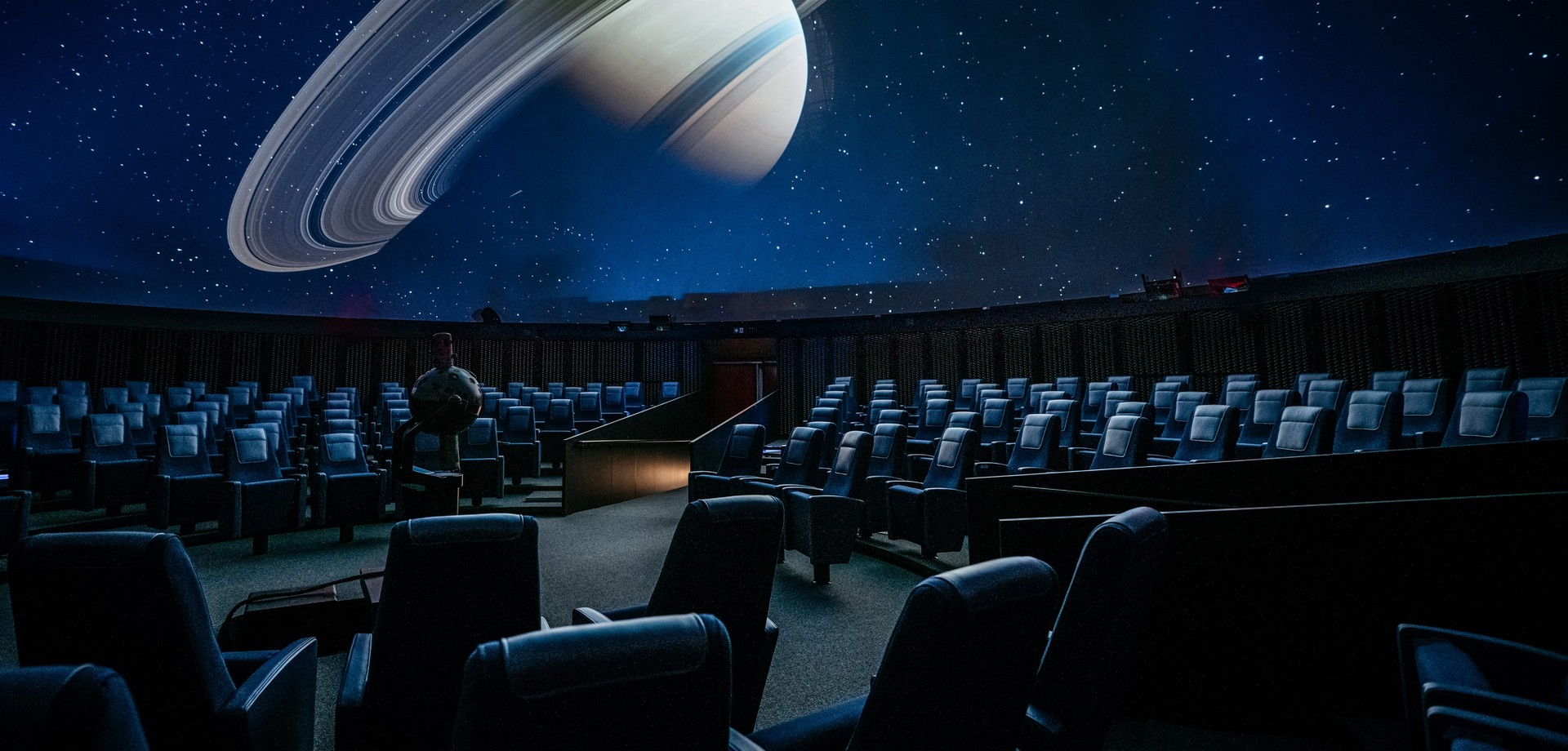 Sitzreihen mit Sesseln und dem runden Sternenprojektionsgerät im Vordergrund. Dahinter ist der Planet Saturn an der Planetariumskuppel zu sehen.