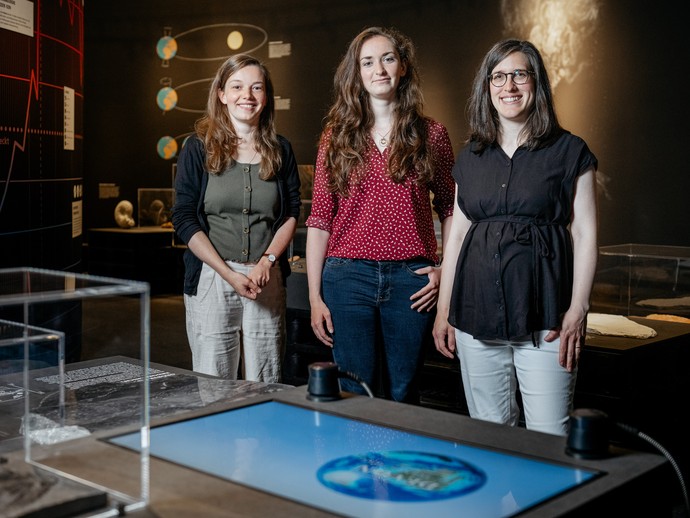 Die drei Ausstellungsmacherinnen stehe in der Ausstellung vor einer Objektinsel mit einem Film und Fossilien. (vergrößerte Bildansicht wird geöffnet)