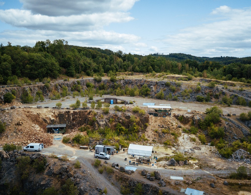 Foto der Grabungsstätte in Balve: Steinbruch mit vereinzelten Sträuchern, Transportfahrzeugen und aufgeschlagenen Zelten.