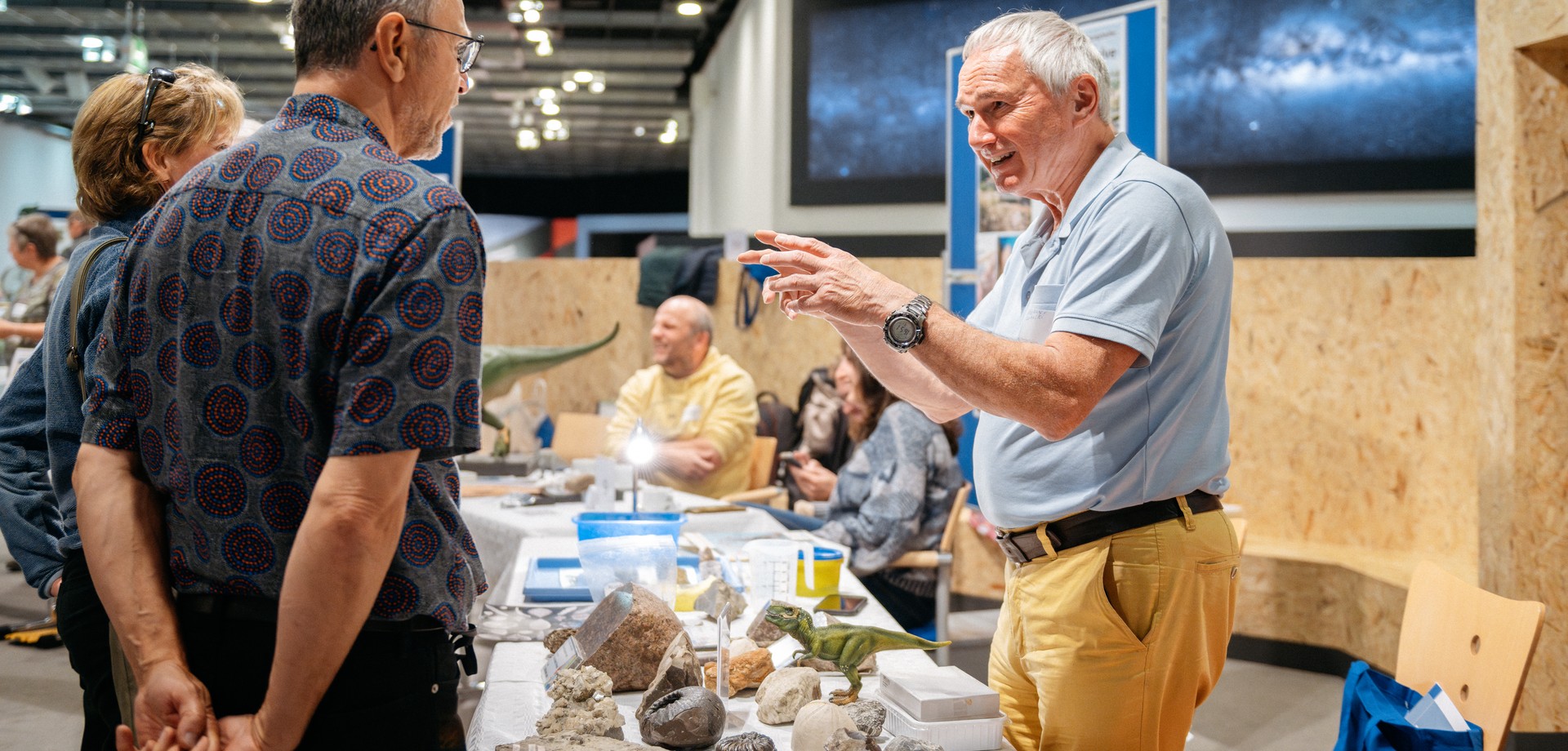 Ein Man steht hinter einem Tisch, auf dem Fossilien liegen, links und rechts neben dem Tisch stehen Menschengruppen. Eine Frau steht an dem Tisch.