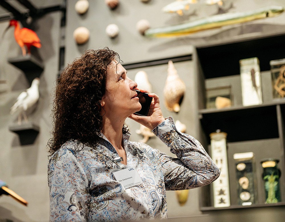 Eine Museumspädagogin steht in der Gene-Ausstellung vor der Vielfaltswand mit vielen verschiedenen Tieren und telefoniert.