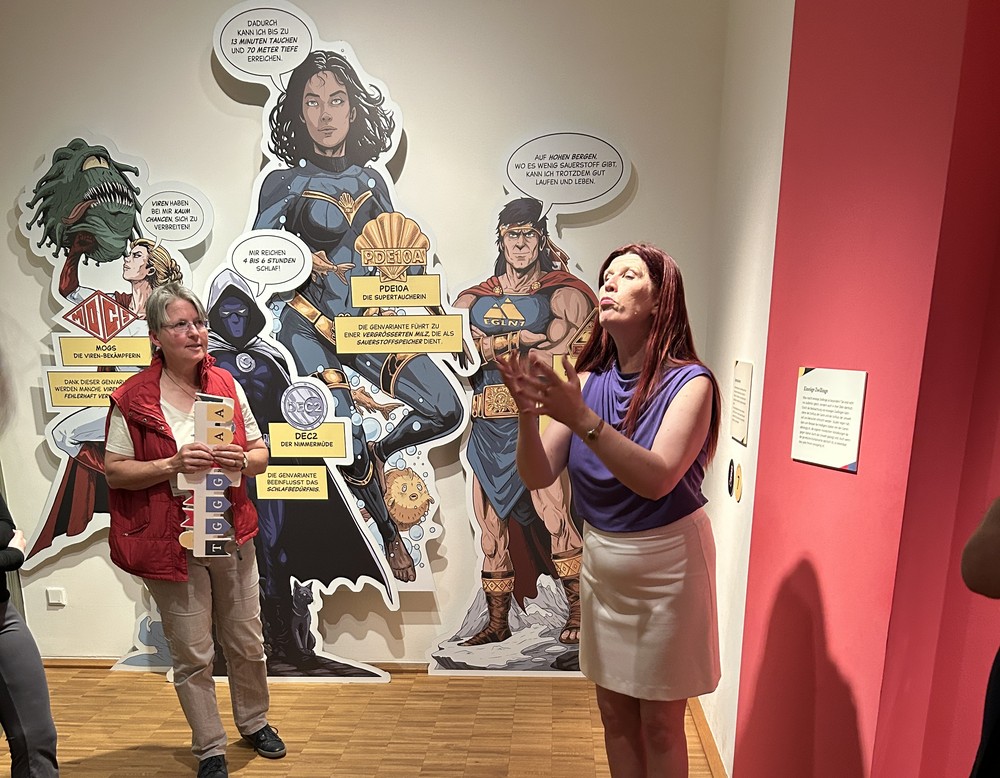 Eine Museumspädagogin steht in der Gene-Ausstellung und erklärt ein Schaubild, neben ihr eine Frau, die in deutscher Gebärdensprache übersetzt.